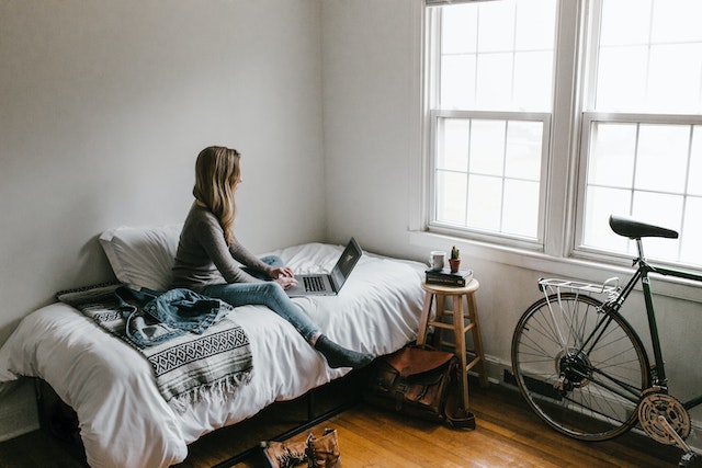 a person typing on a laptop on a bed next to a stool and bike